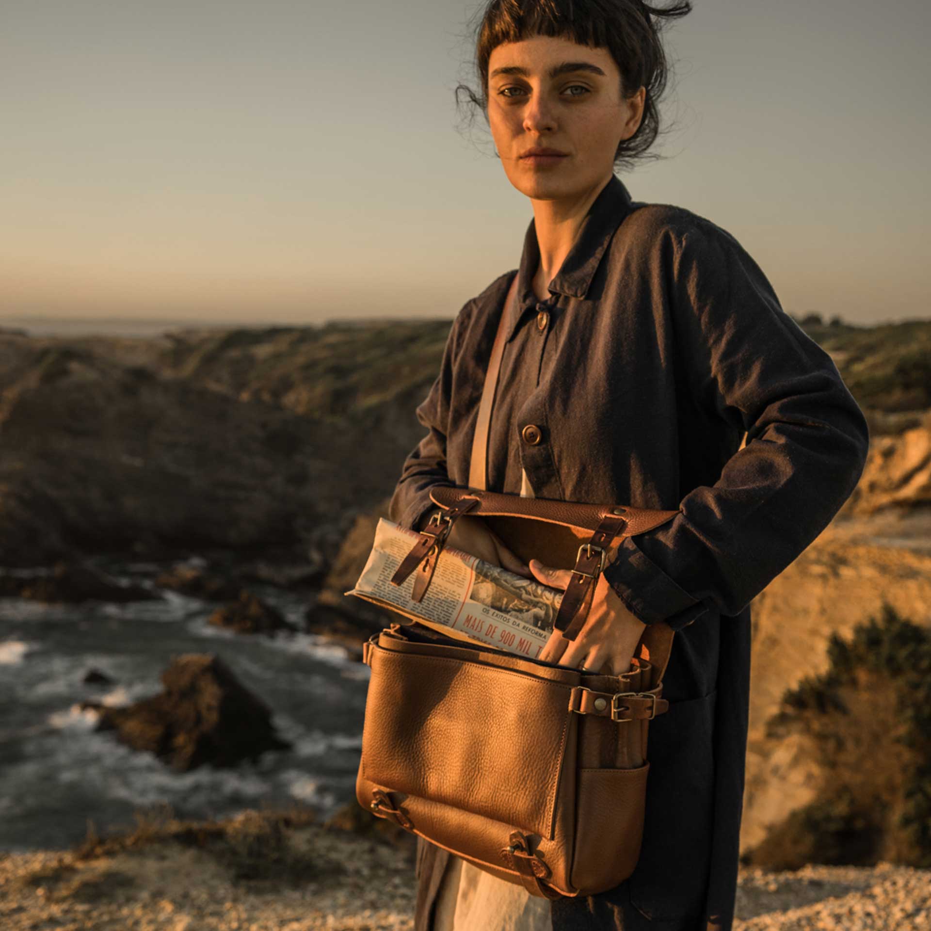 Une femme porte le sac en cuir "Eclair" en bandouliere, le rabat est ouvert et elle y glisse un journal.