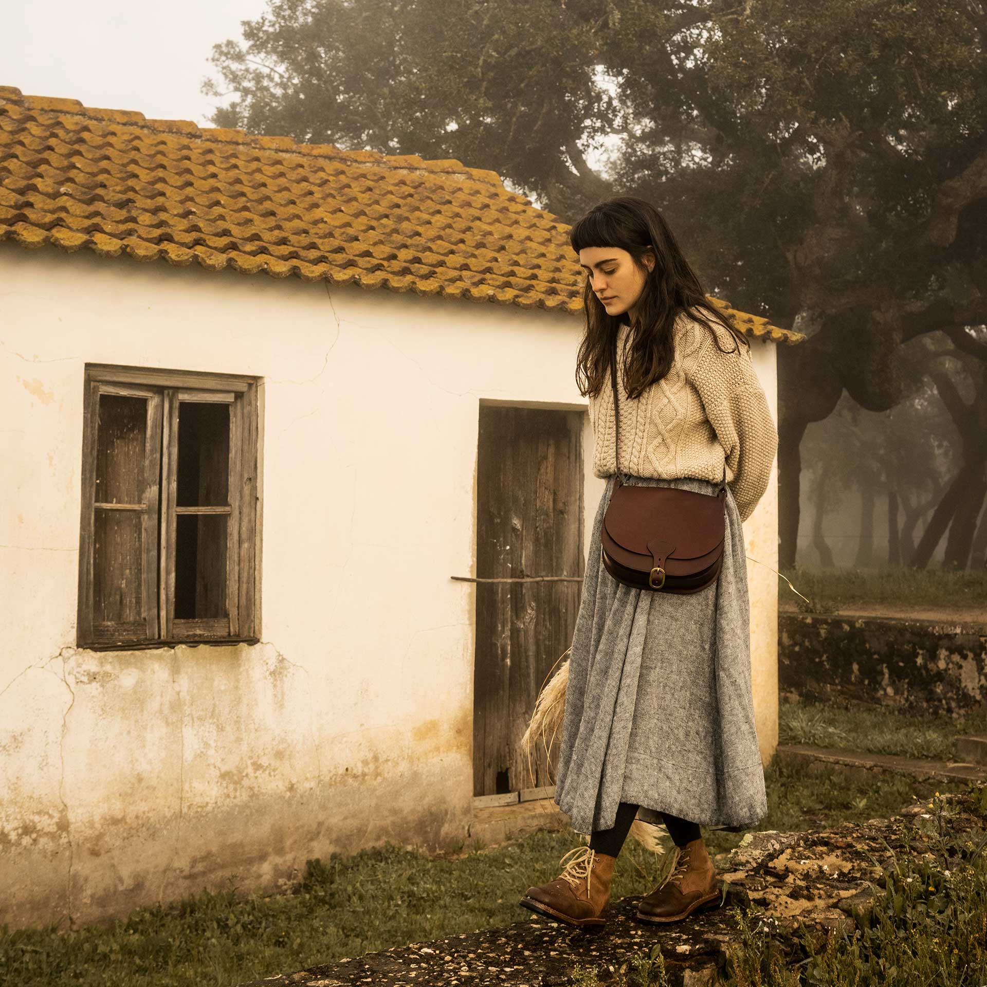 Une femme porte en bandouli&egrave;re une besace en cuir devant une vieille maison