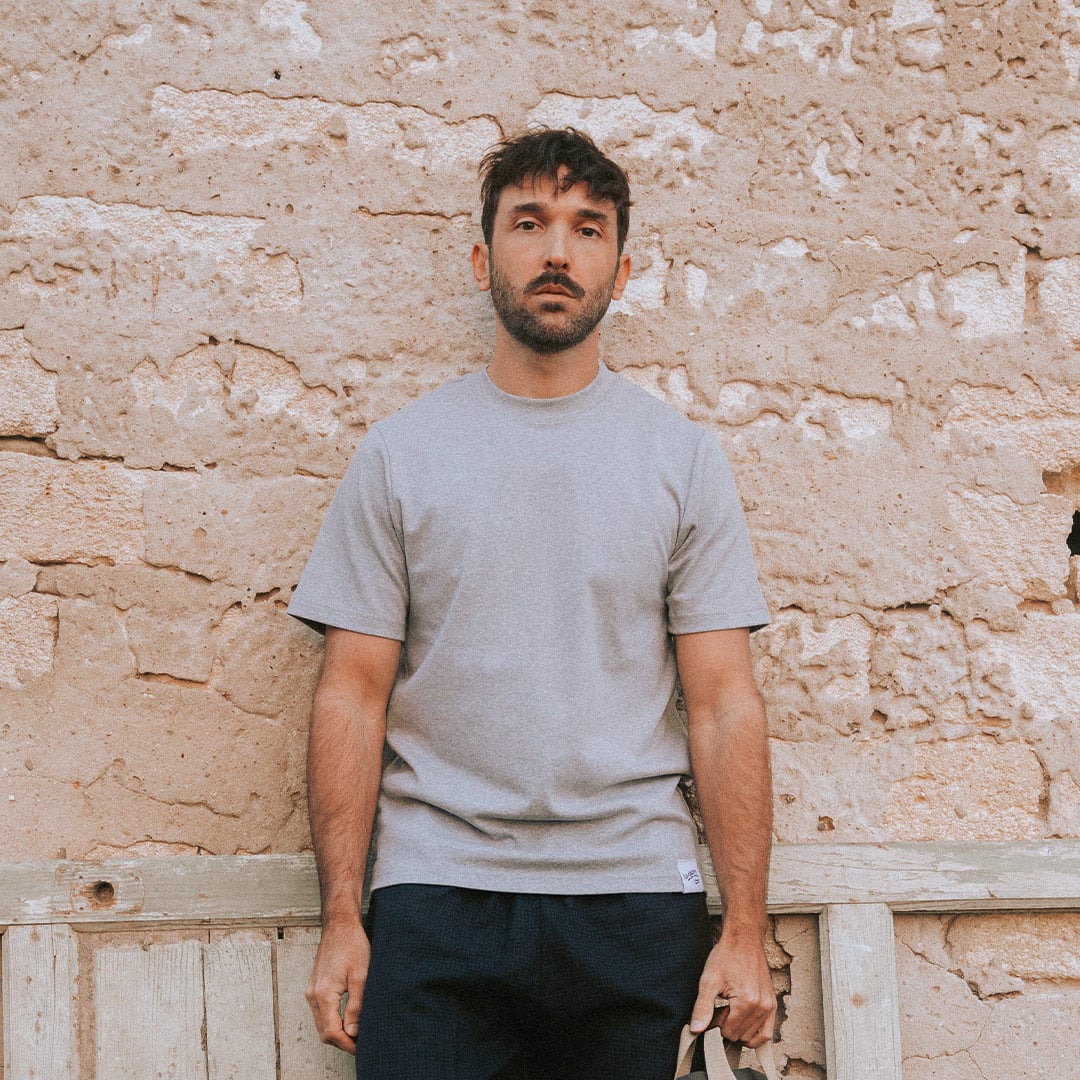 Un hombre apoyado contra una pared lleva una camiseta gris «Made in France».