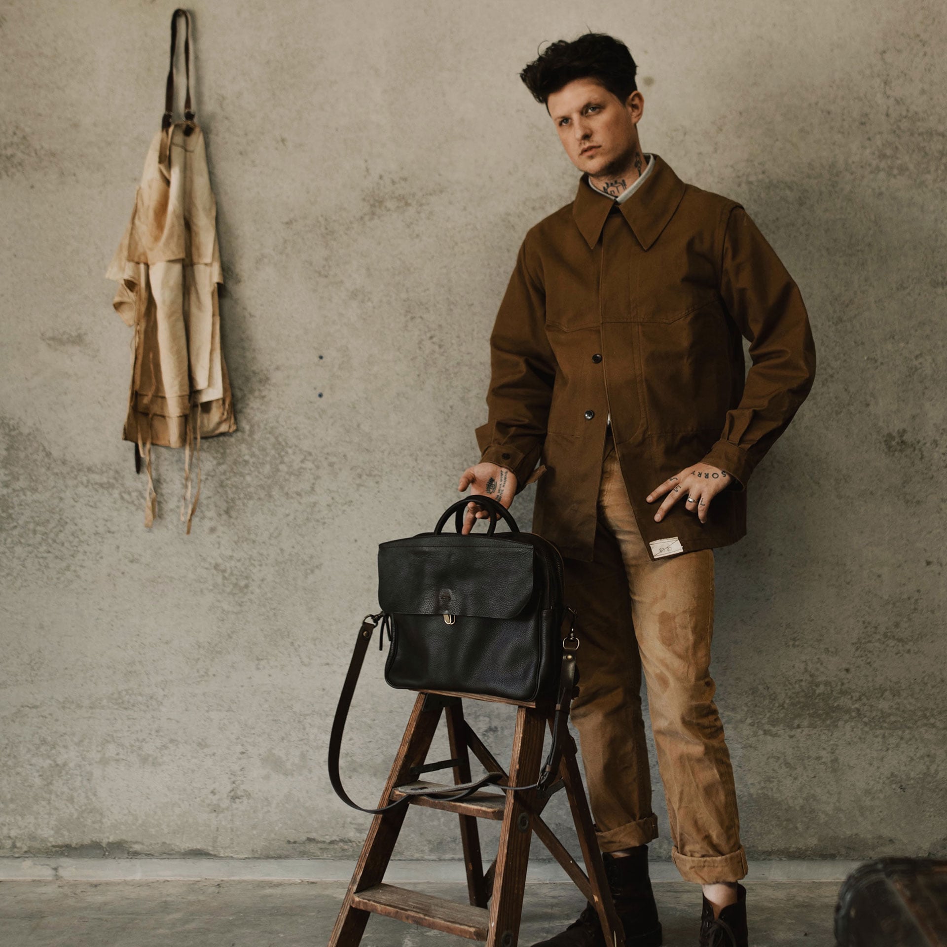 Un homme au style workwear se porte a cot&eacute; de son sac en cuir