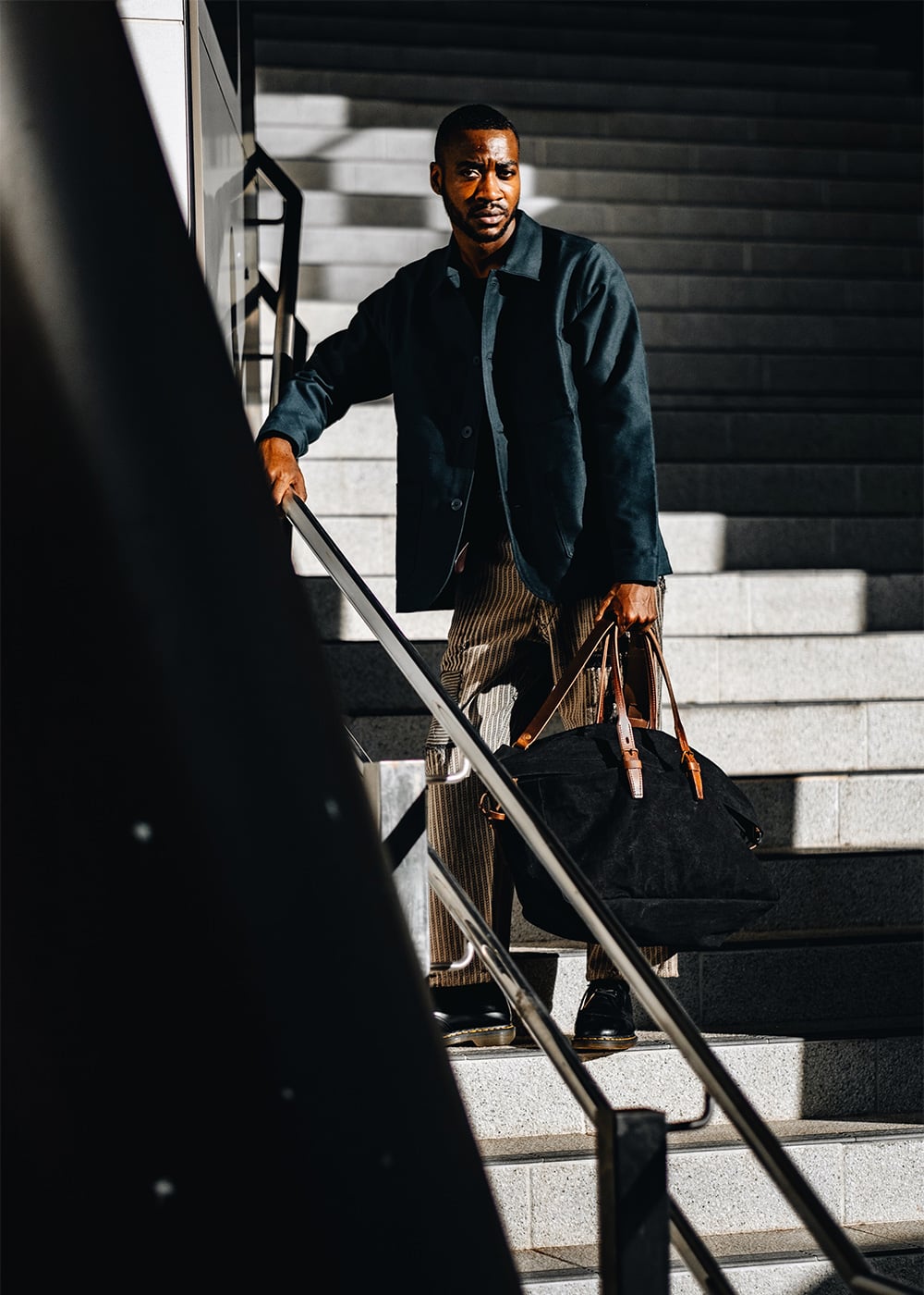 Un homme pose sur un escalier et tient a bout de bras un sac de voyage en toile
