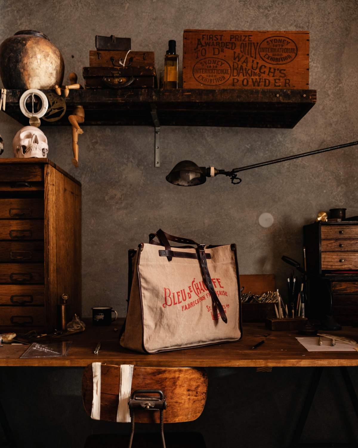 a canvas tote bag sits on a vintage wooden desk, inscribed &ldquo;BLEU DE CHAUFFE - FABRICATION FRANCAISE - St-Georges-de-Luzencon&rdquo;.