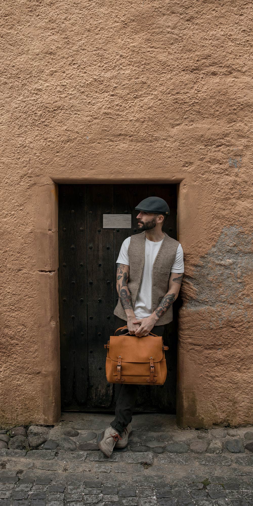 A man in front of a wall carries his leather satchel at arm's length.