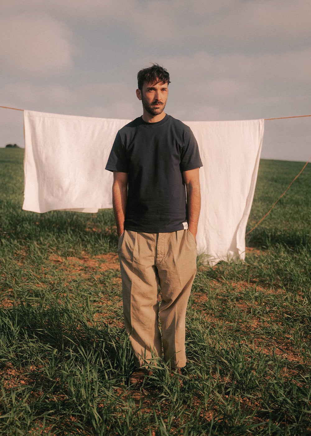 Un hombre en un prado, delante de unas sábanas blancas, lleva una camiseta azul de trabajo.