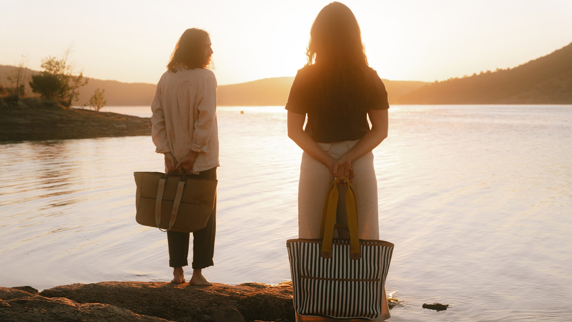 A man and a woman are carrying canvas tote bags in front of Lake Salagou at sunset.